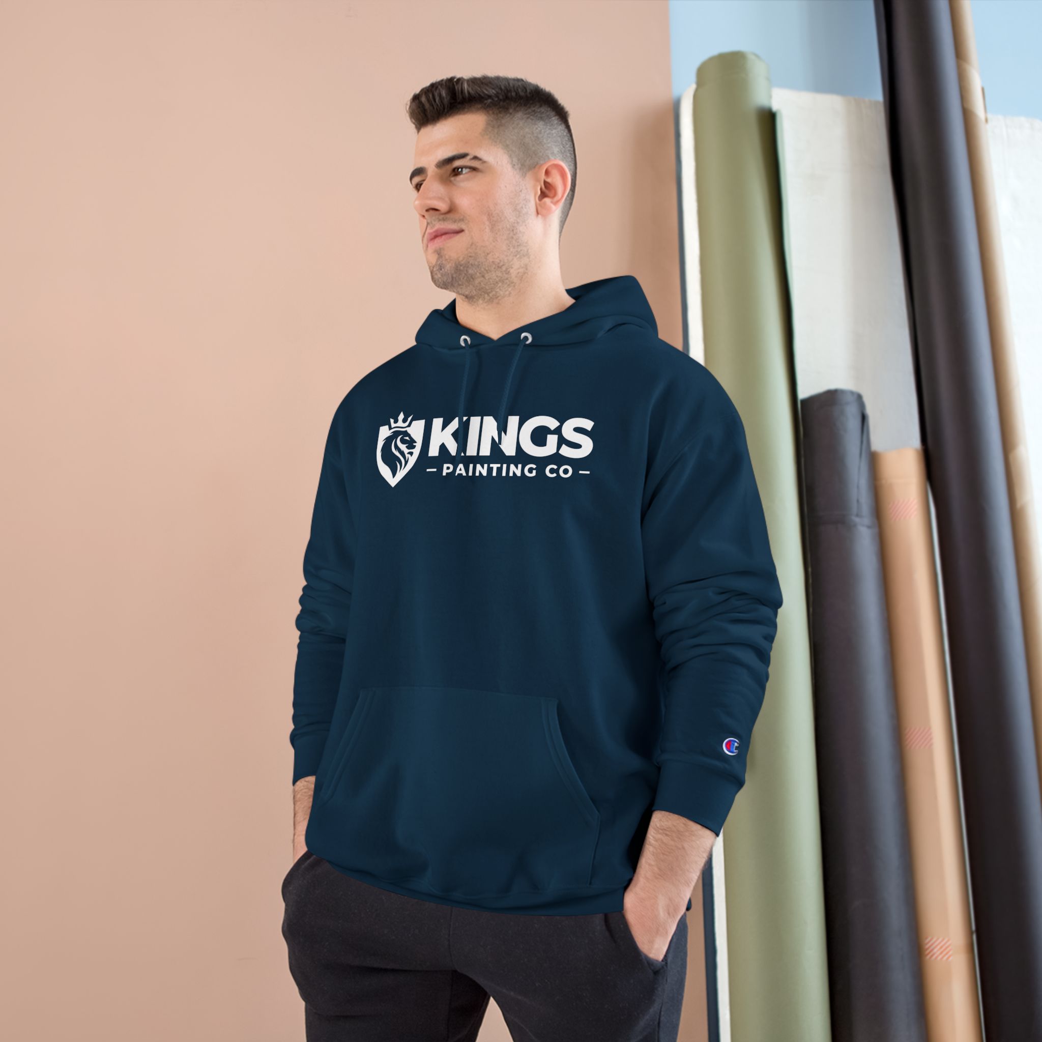 A man wearing the Kings Painting Co. Logo Hoodie by Champion® stands indoors beside rolls of fabric or paper, looking to the side and smiling.