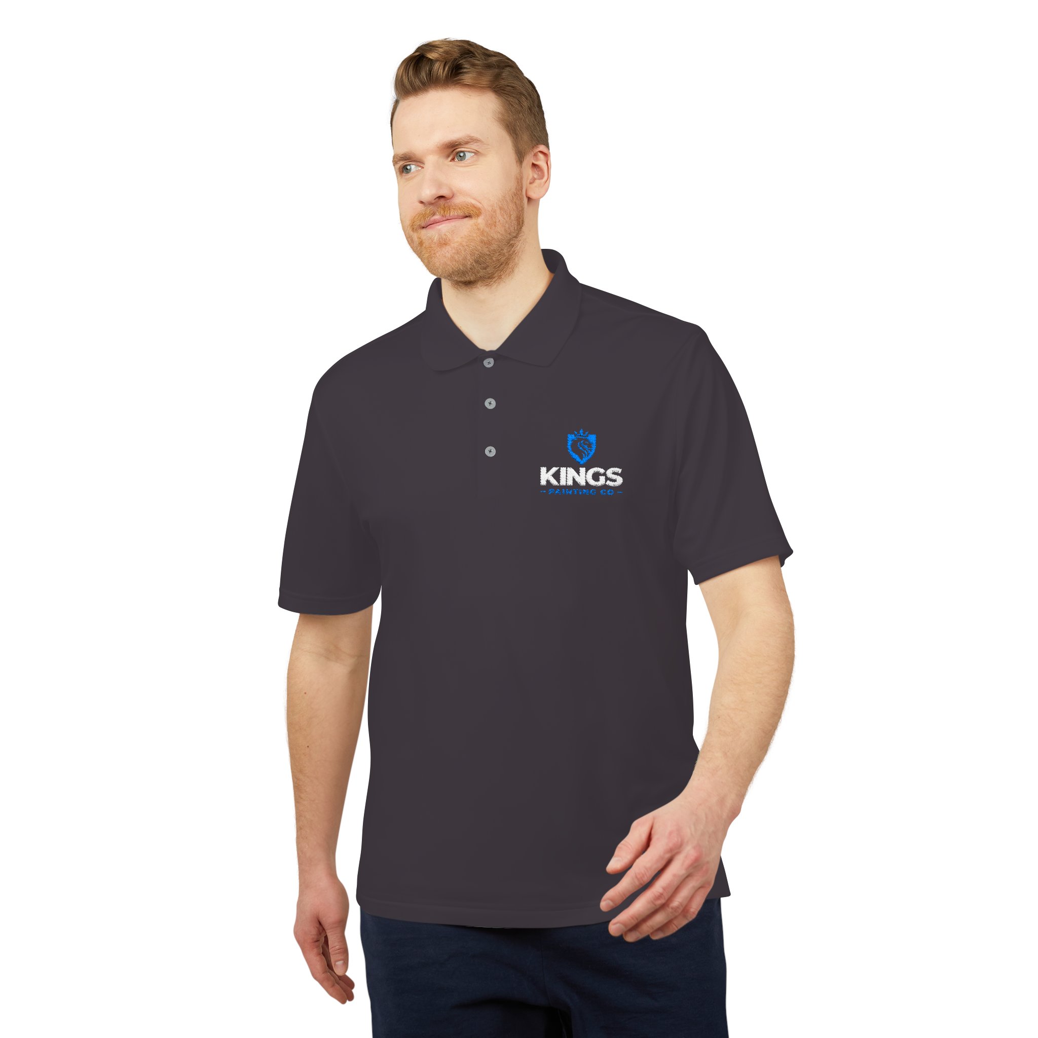 A man with light hair is wearing the Kings Painting Co. Performance Polo by Under Armour®, which features the Kings logo and a blue lion emblem on the left chest; he stands looking slightly to the side with a neutral expression.