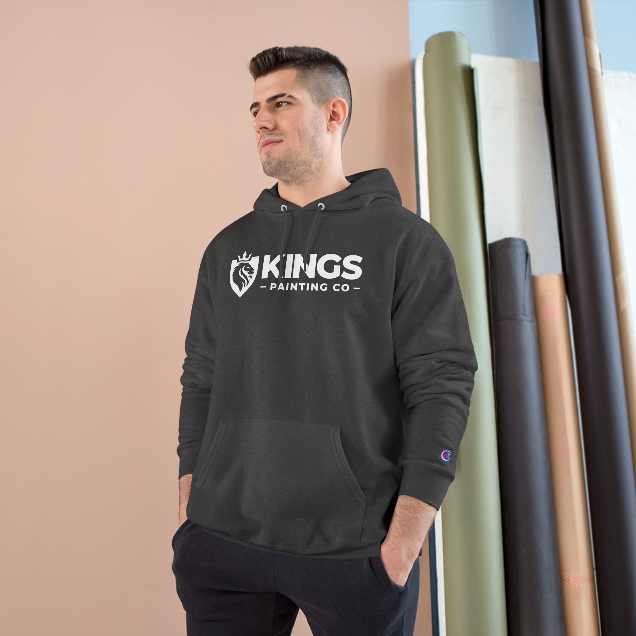 A man wearing the Kings Painting Co. Logo Hoodie by Champion® stands indoors near rolls of colored paper, looking to the side and smiling slightly.