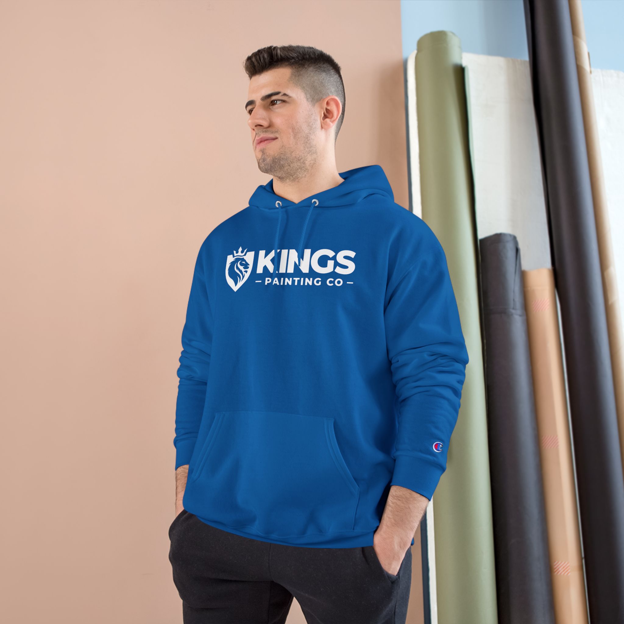 A man stands facing left with his hand in his pocket, wearing the Kings Painting Co. Logo Hoodie by Champion®, in blue. Rolled materials are stacked behind him against a beige and white wall.
